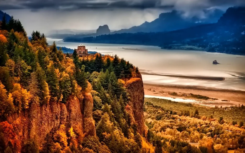 A scenic view shows a historic building perched on a cliff surrounded by a vibrant mix of autumn-colored trees. Below, a wide river winds through a lush valley, with misty mountains forming a dramatic backdrop under a cloudy sky.