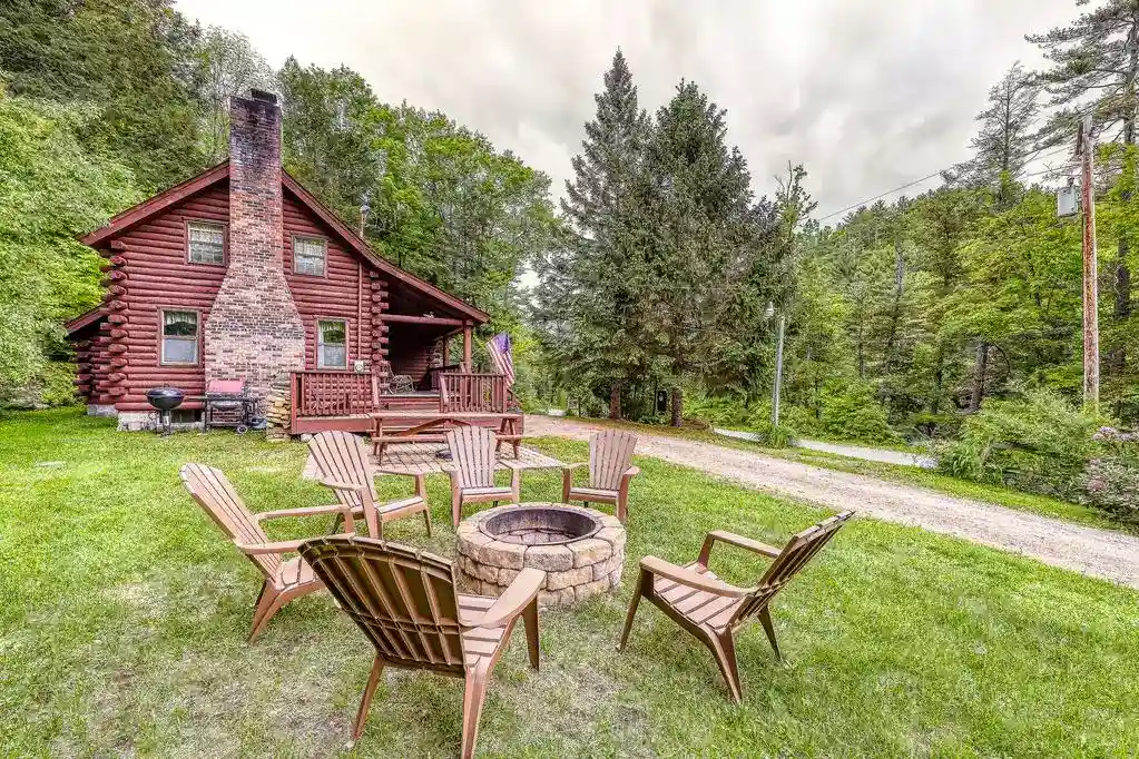 A rustic log cabin with a brick chimney nestled in a lush forested area. In the grassy yard, wooden chairs surround a stone fire pit. A gravel path leads away from the cabin, surrounded by tall trees and greenery. Soft light filters through the overcast sky, creating a peaceful atmosphere.