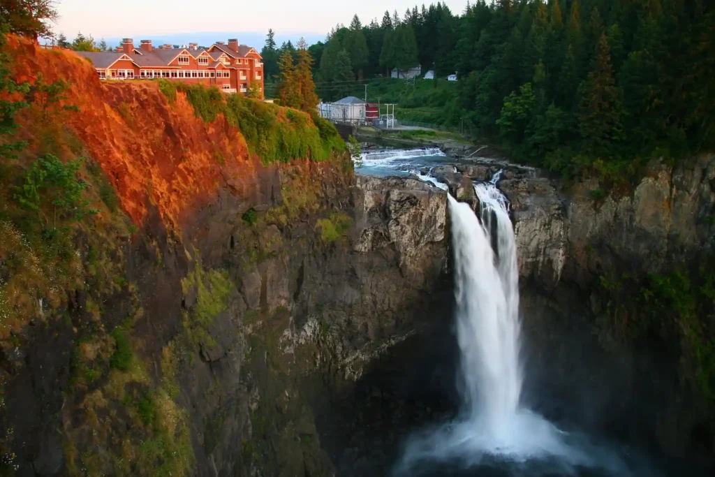 A picturesque view of a waterfall cascading from a cliff into a pool below, surrounded by lush green forest. A large lodge, bathed in warm sunset light, perches atop the neighboring cliff, overlooking the scenic waterfall and river. A small structure sits near the water below.