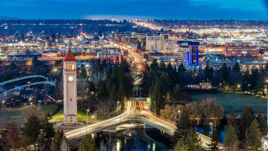 An evening aerial view of a bustling cityscape featuring a prominent clock tower with a red roof, surrounded by illuminated buildings and winding streets. A bridge and a river can be seen in the foreground, leading into a park with scattered trees.