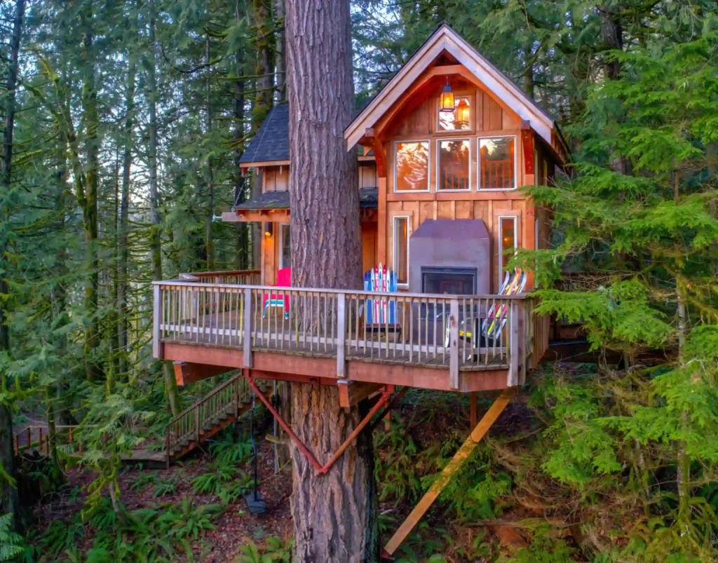 A treehouse nestled in a lush forest features a wooden deck with colorful chairs. The structure has large windows, a sloping roof, and is built around a sturdy tree trunk. Vibrant greenery surrounds the elevated, cozy retreat.