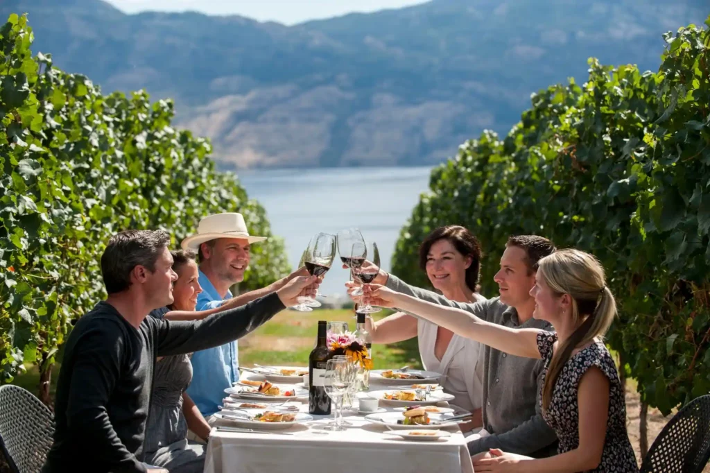 A group of six people sits around a table set with food and wine, toasting with glasses of red. The table is nestled between lush green grapevines, with mountains and a body of water visible in the background, suggesting a vineyard setting.