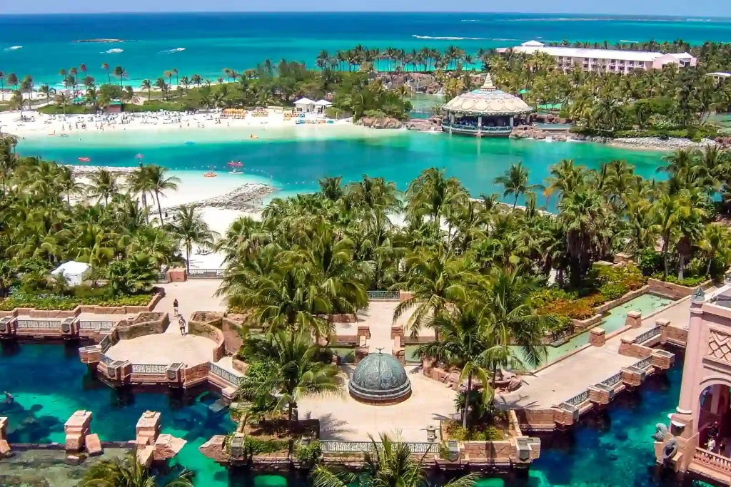 A stunning view of a tropical resort in the Bahamas features lush palm trees, turquoise water, white sandy beaches, and several thatched-roof structures. The scene is bordered by vibrant greenery, with an array of pools and pathways weaving through the resort area.