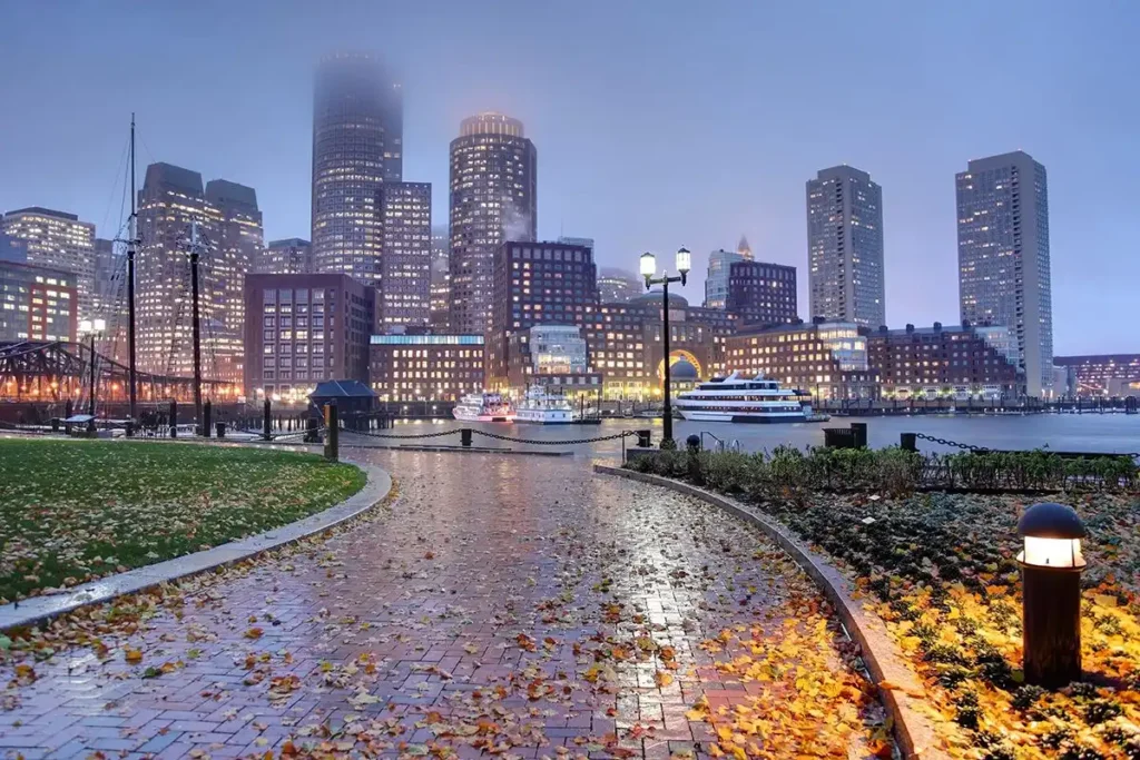 A foggy evening cityscape in Boston showcasing a waterfront park with a brick pathway and lush greenery. Tall buildings with illuminated windows stand in the background, and boats are visible docked near the shore. Lamps along the path cast a warm glow over this Massachusetts haven.