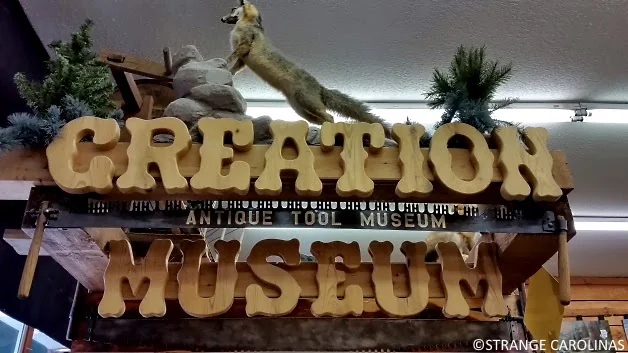 A sign that reads "CREATION MUSEUM" with "ANTIQUE TOOL MUSEUM" written below it. The sign is surrounded by small pine trees, and a taxidermy fox is positioned at the top of the sign. The background appears to be an indoor setting.