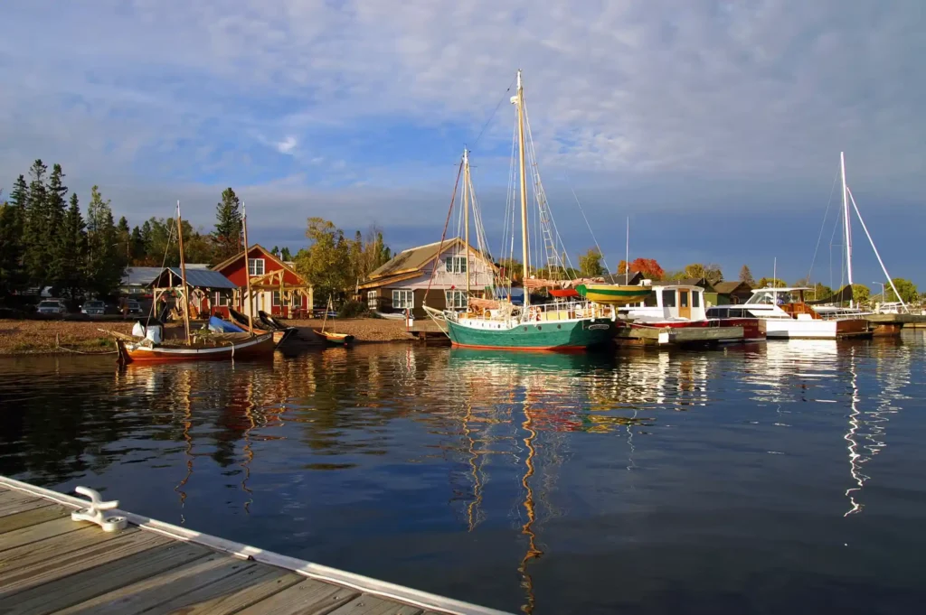 Calm harbor scene with several boats docked along a pier in Grand Marais, Minnesota. A green sailboat is prominent, with a red building and pine trees in the background. The water reflects the boats and sky, which is partly cloudy. A wooden dock is visible in the foreground.