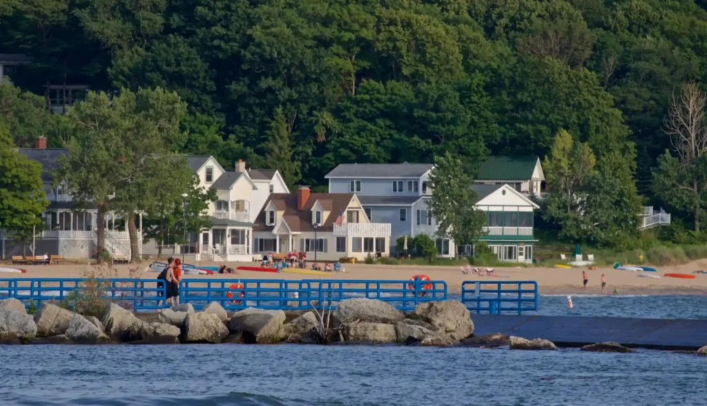 Several houses sit along a sandy beach in Holland, Michigan, surrounded by lush greenery. A few people stand on a blue pier that extends over the water, with large rocks at the shoreline. The calm water in the foreground contrasts with the vibrant colors of the beach scene.