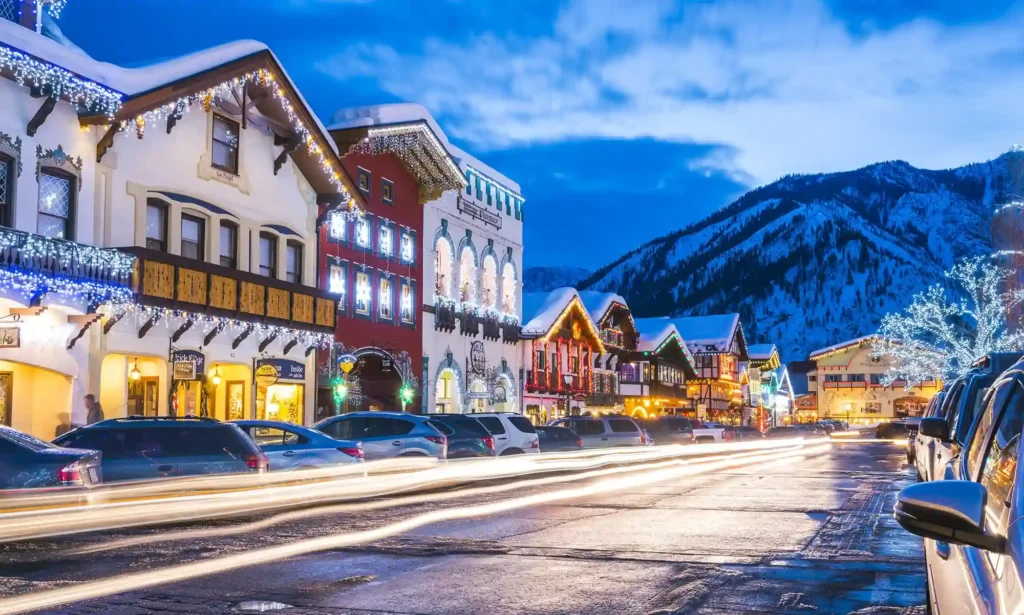 A festive street scene in the small town of Leavenworth, Washington, with European-style buildings adorned with holiday lights. Snow-capped mountains rise in the background under a twilight sky. Car light trails blur in the foreground, indicating motion on the street.