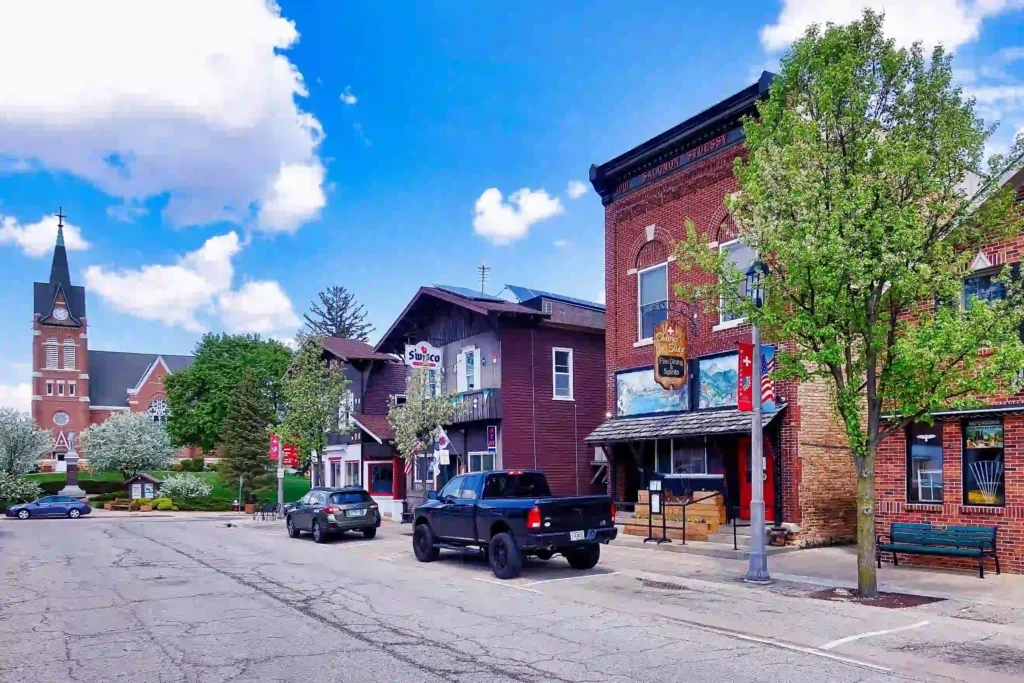 A small-town street in New Glarus, Wisconsin, lined with vintage brick and wooden buildings under a bright blue sky with scattered clouds. Parked cars are along the street, and a few trees are in bloom. A tall church steeple is visible in the background on the left.