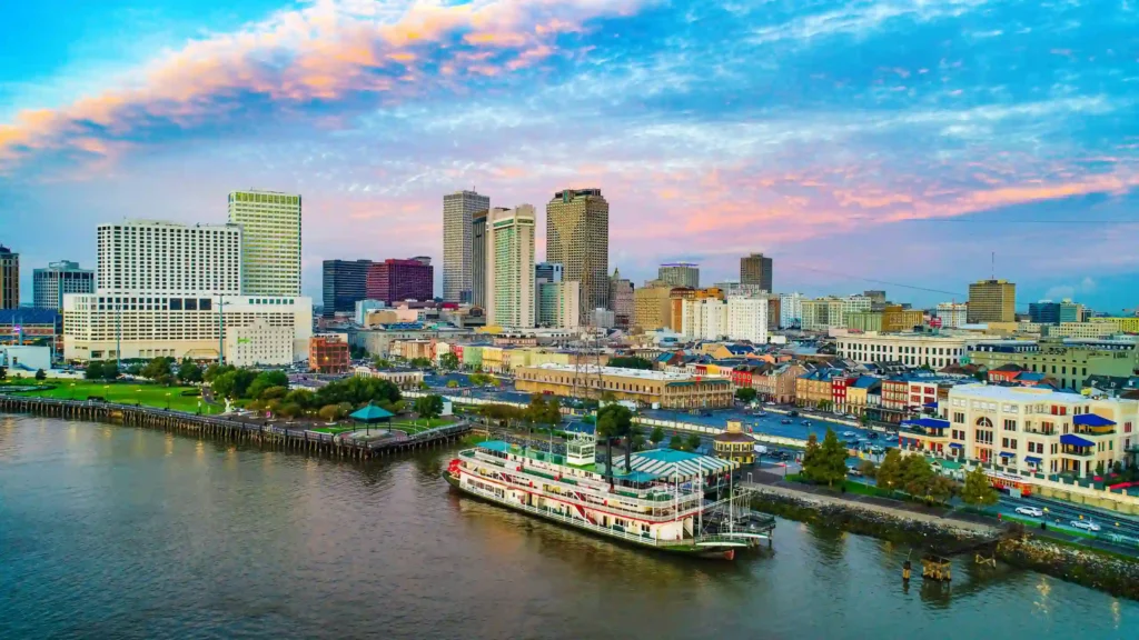 A vibrant cityscape of New Orleans, Louisiana with tall buildings against a colorful sunset sky. The Mississippi River is in the foreground, featuring a classic paddlewheel steamboat docked next to a waterfront park and historic district buildings.