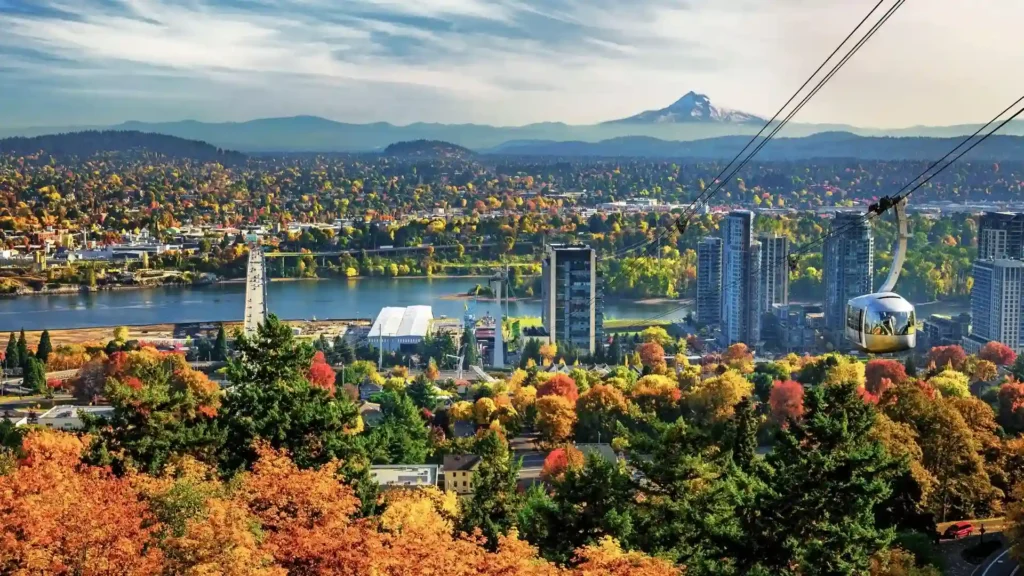 Show thumbnail preview A scenic view of Portland, Oregon with a mix of tall buildings and lush, colorful autumn trees. In the background, mountains with blue skies and scattered clouds are visible. A cable car moves along a cable over the greenery, adding to the picturesque landscape.