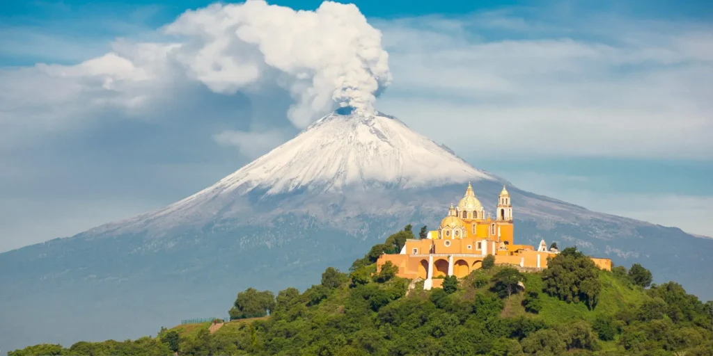 A yellow church sits atop a green hill, with trees surrounding it. In the background, a snow-capped volcano emits a column of white smoke into the blue sky. The scene in Puebla, Mexico captures a striking contrast between human architecture and natural beauty.