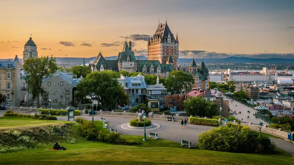 The image shows a scenic view of Quebec City, Canada, with the iconic Château Frontenac standing prominently in the center. The surrounding area features historic buildings, lush greenery, and pathways. The sky is painted with hues of orange and pink during sunset.