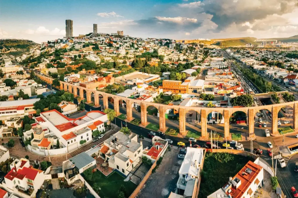 Aerial view of Querétaro, Mexico, showcasing the iconic aqueduct with multiple arches running through the city. Residential and commercial buildings are spread out, with a hill in the background featuring modern high-rises. The sky is partly cloudy.