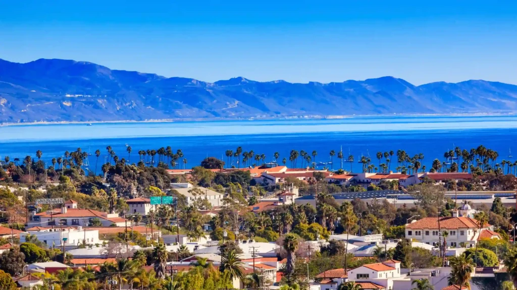 A coastal city in California with red-roofed buildings and palm trees, set against a backdrop of calm blue ocean waters and distant mountain ranges under a clear blue sky, Santa Barbara is truly picturesque.