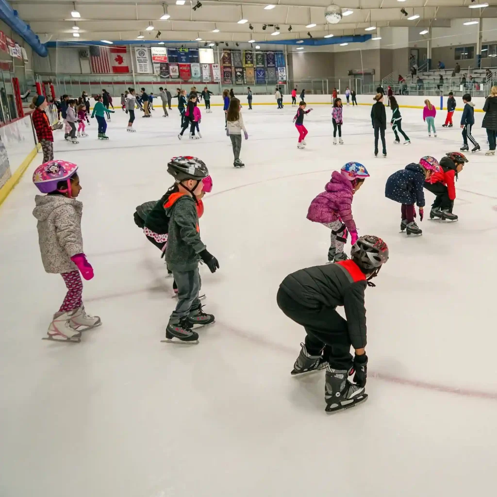 Children and adults are ice skating in a large indoor rink at Ashburn Ice House. The children in the foreground wear colorful helmets and winter clothing, practicing their skating posture. The background features banners and spectators watching from the sidelines.