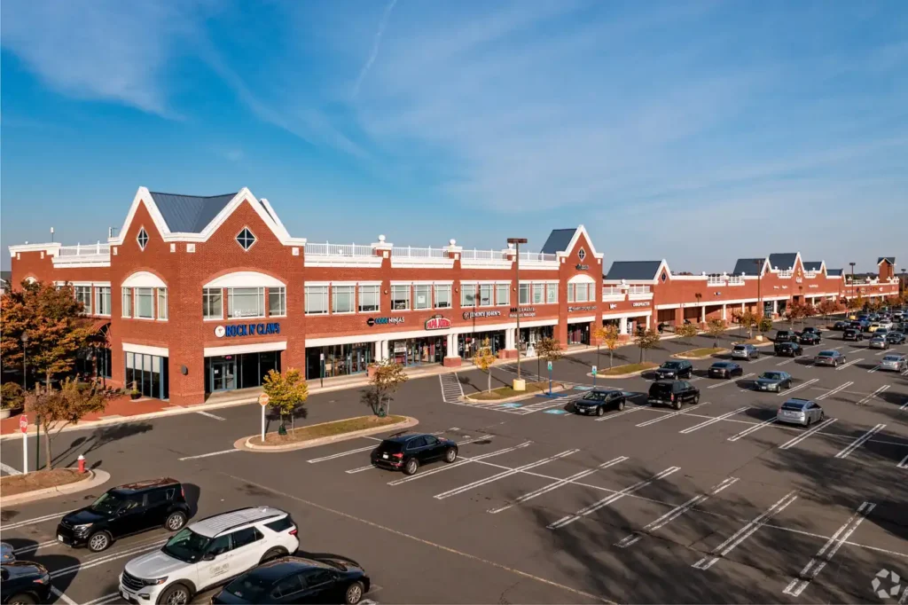 Ashburn Village Shopping Center is a large suburban shopping center with multiple brick storefronts, including a restaurant and various retail stores. The parking lot in front is partly full with a mix of vehicles parked and driving, while a bright blue sky is seen overhead.