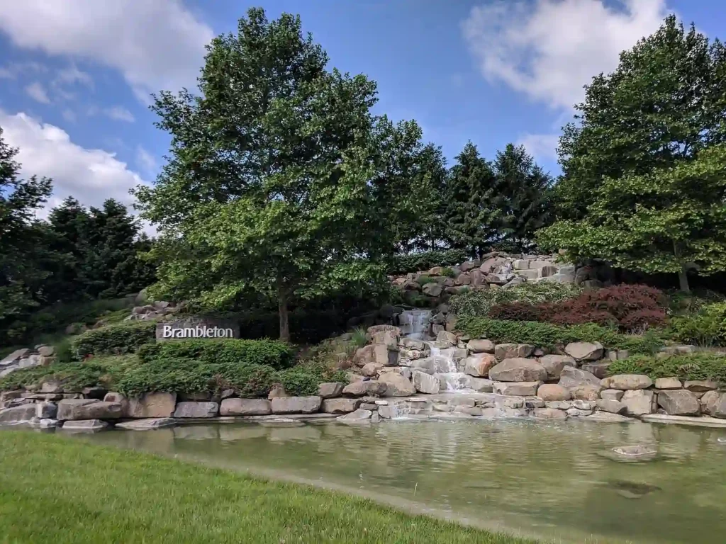 A stone waterfall with trees and a sign that reads "Brambleton" welcomes visitors to Brambleton Regional Park, nestled in a lush landscape under a clear blue sky.