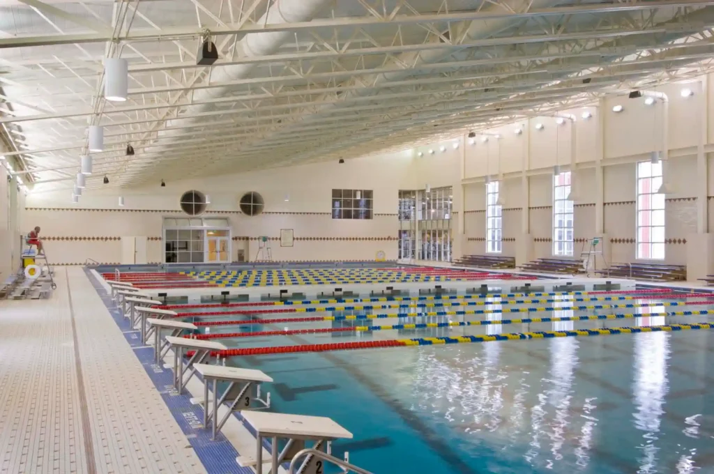 A spacious indoor swimming pool at Claude Moore Recreation Center features multiple lanes separated by red, yellow, and blue lane dividers. Starting blocks are positioned at one end of the pool. The area is surrounded by large windows, white walls, and a high ceiling with exposed beams.