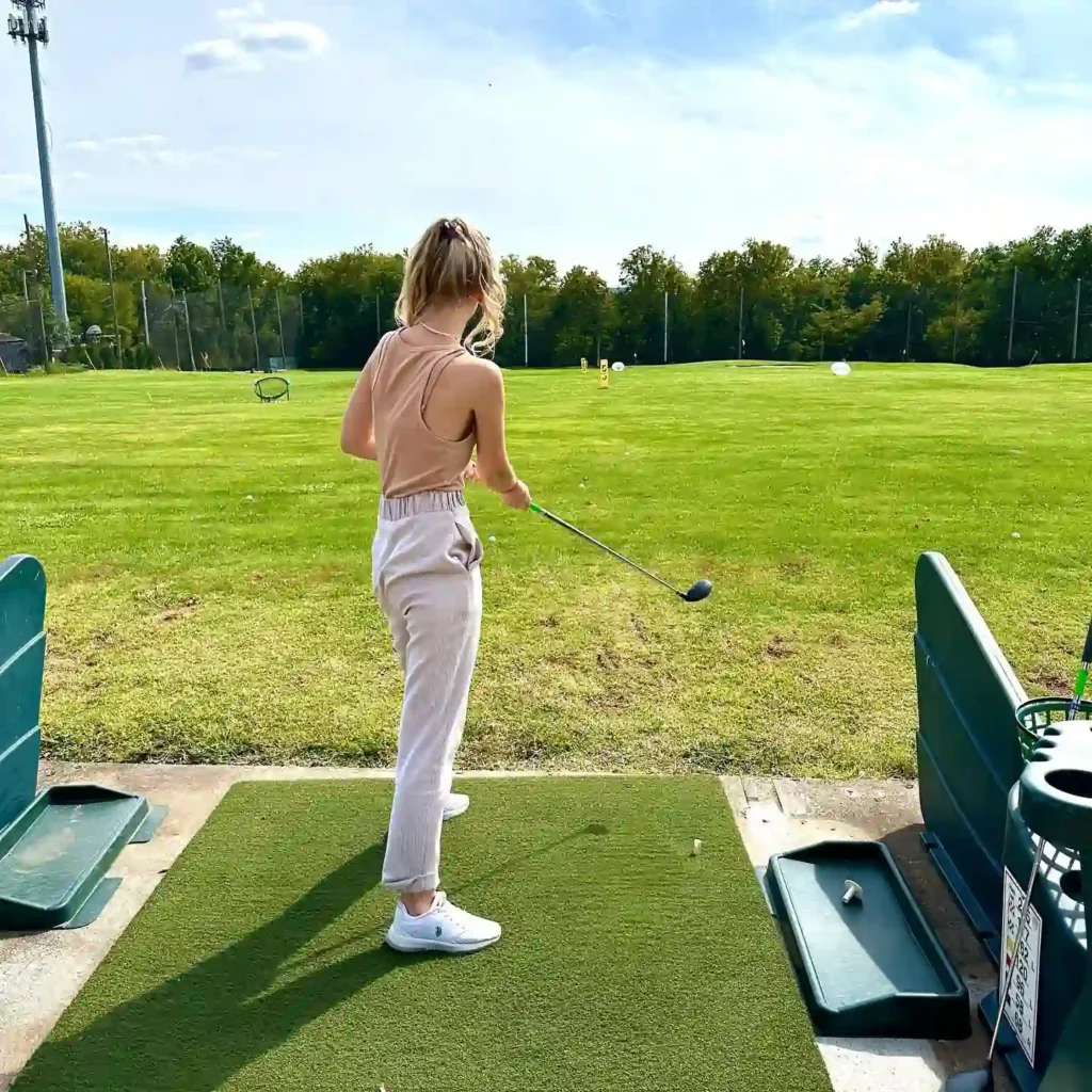 Person in casual attire preparing to hit a golf ball at the Dulles Golf Center & Sports Park driving range on a sunny day.