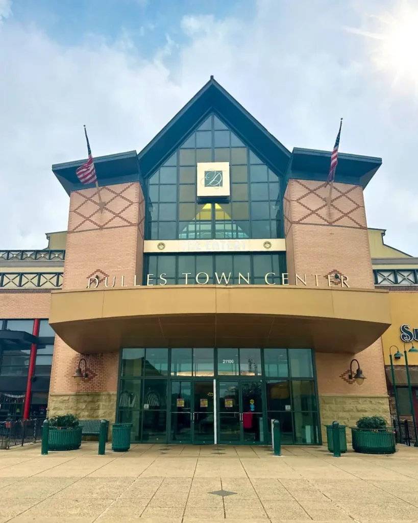 Entrance to Dulles Town Center mall with glass doors, U.S. flags, and brick facade on a cloudy day, showcasing a welcoming atmosphere for shoppers.