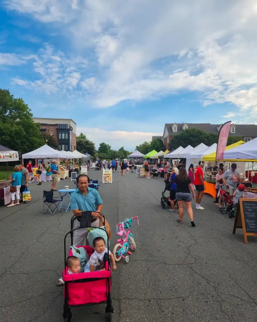 A person pushes a pink double stroller with two young children through the bustling EatLoco Farmer's Market. The street is lined with white vendor tents and brimming with people. The sky is partly cloudy, adding to the vibrant atmosphere of the market.