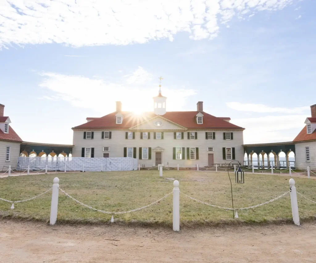 A large historic house with a red roof, white exterior, and multiple chimneys stands under a partly cloudy sky, surrounded by a lawn and white posts connected by chains—reminiscent of George Washington’s Mount Vernon.