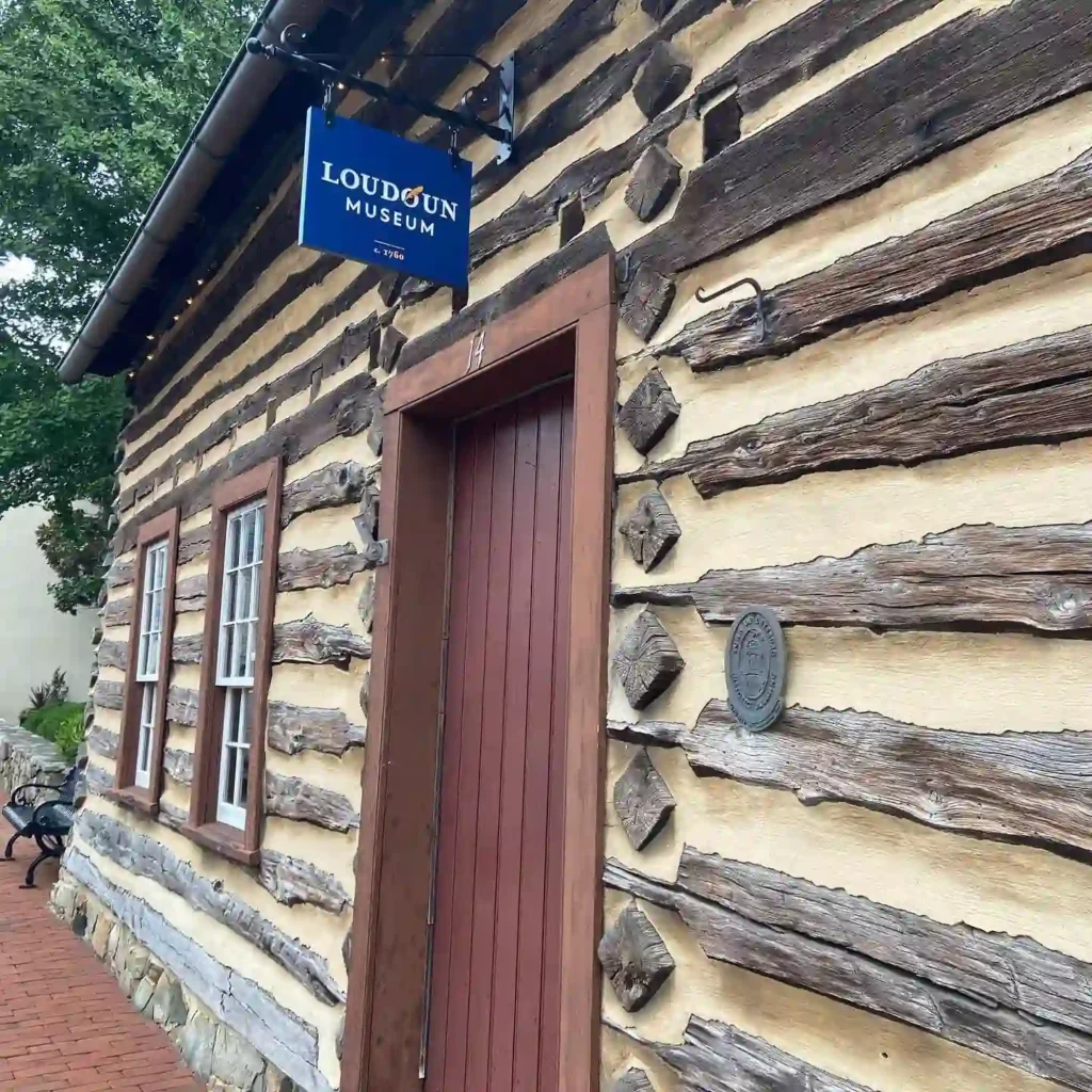 A log cabin with a wooden door and windows, labeled "Loudoun Museum" on a sign above. The exterior showcases weathered wooden logs and a historical plaque near the entrance.