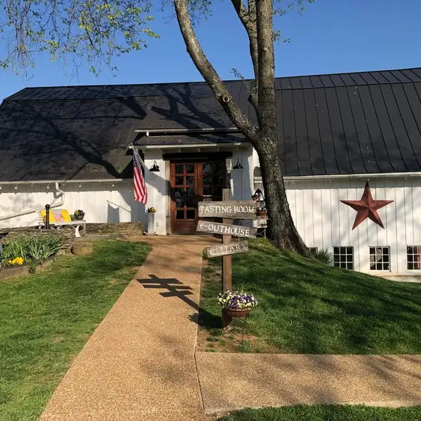 A rustic building with a "Tasting Room" sign, American flag, brown door, red star decoration, and a tree in the yard stands charmingly at The Barns at Hamilton Station Vineyards.