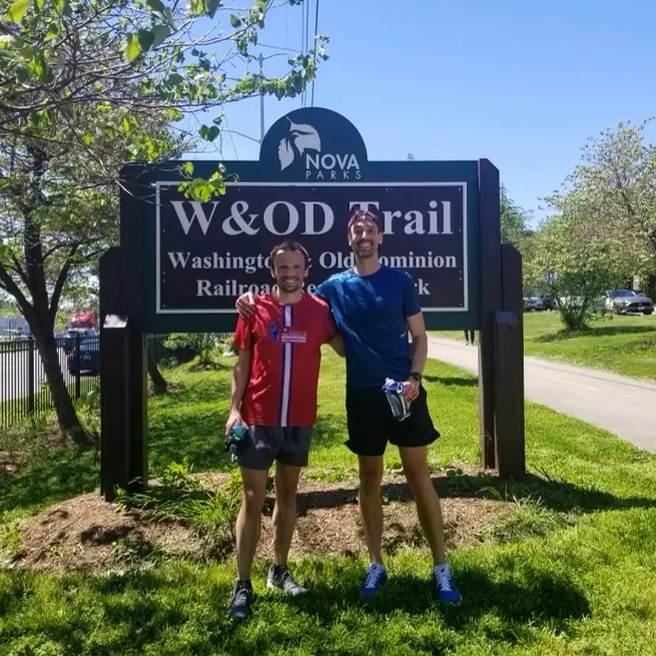 Two people in running attire stand smiling in front of a Washington & Old Dominion Trail sign on a sunny day.