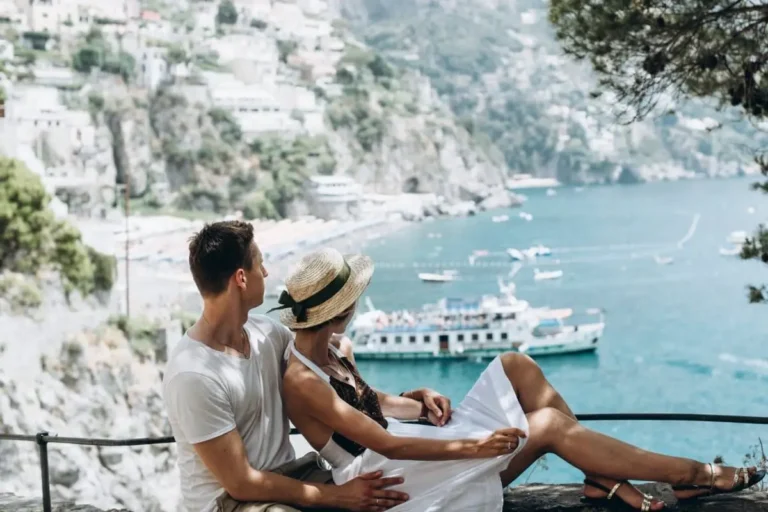 A couple sits on a ledge which is one of Best Honeymoon Destinations On A Budget, overlooking a scenic coastal view with boats on the water. They are dressed in summer clothing; shes wearing a straw hat. The background features cliffs, houses, and a vibrant blue sea.