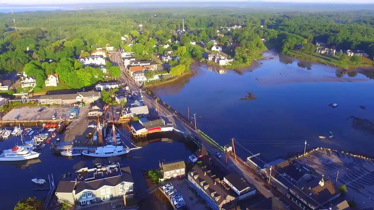 Aerial view of a coastal town with lush green vegetation, a river, and small boats docked at a marina. Roads and bridges connect the residential and commercial buildings, creating a picturesque community surrounded by natural beauty.